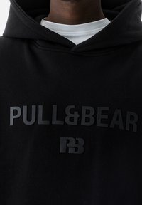 Personne portant un sweat à capuche noir avec les logos "PULL&BEAR" et "PB" en lettres noires en relief sur un t-shirt blanc à col rond en dessous.