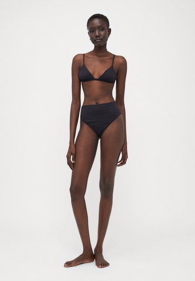 Black bikini set featuring a triangular top with thin straps and high-waisted bottom, both made from a smooth fabric. Model stands barefoot.