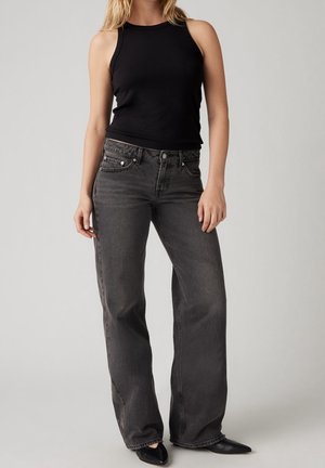 Kvinde iført sort ærmeløs top, sorte, løstsiddende jeans med højtaljede og spidse sorte sko, står foran en ensfarvet baggrund.