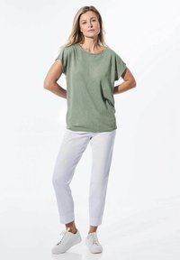 Hellgrünes, kurzärmliges Oberteil in lockerer Passform; kombiniert mit weißen, knöchellangen Hosen. Die Schuhe sind weiße Sneaker mit Schnürsenkeln. Minimalistisches Design.