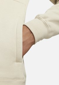Beige sweatshirt med texturerad tyg. Har en känguruficka och ribbade muddkanter. Närbild på arm med hand i fickan.