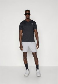 Juoda trumpomis rankovėmis marškinėliai su logotipu, pilkos kelnės šortai, juodos kojinės ir pilki sportbačiai. Modelis dėvi akinius nuo saulės, stovi prie šviesaus fono.