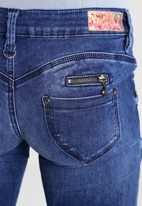 Vaqueros de denim azul oscuro con un bolsillo trasero con cremallera, trabillas para cinturón y una etiqueta de tela floral en la cintura. Textura suave, diseño ajustado.