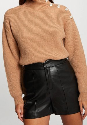 Woman wearing a camel ribbed turtleneck sweater with heart-shaped pearl buttons and high-waisted black leather shorts.