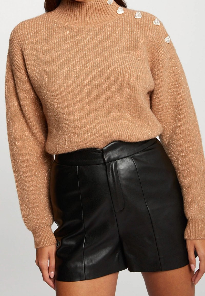 Femme portant un pull à col roulé côtelé couleur camel avec des boutons en perle en forme de cœur et un short en cuir noir taille haute.