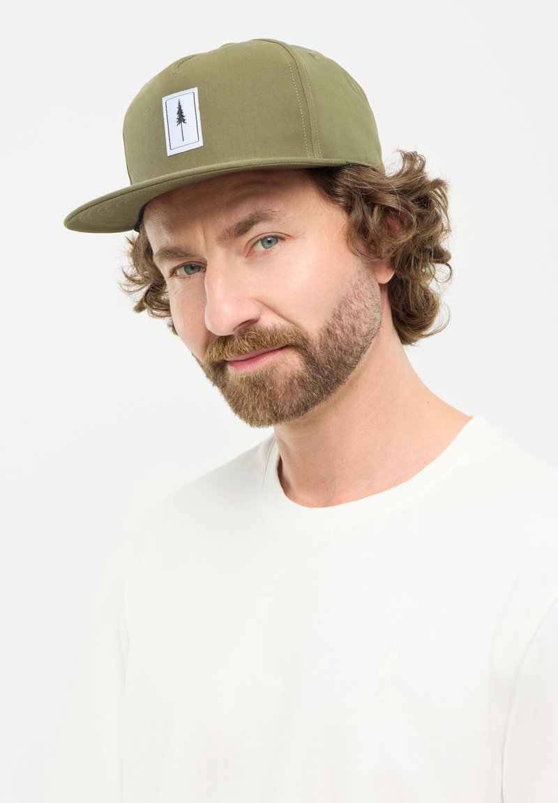Olijfgroene pet met een vlakke rand en een witte patch met een zwart boomontwerp; gedragen met een eenvoudig wit shirt.