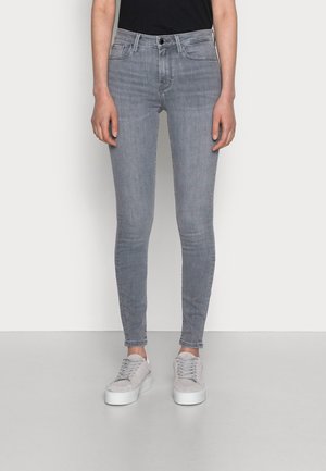 Schmal geschnittene graue Jeans aus Denim, getragen mit hellgrauen Sneakers von einer stehenden Person vor einem schlichten weißen Hintergrund.