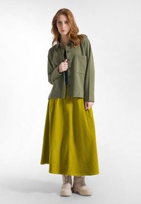 Groene katoenen shirtjasje met een borstzak, gecombineerd met een getexturede gele maxirok en beige veterschoenen.