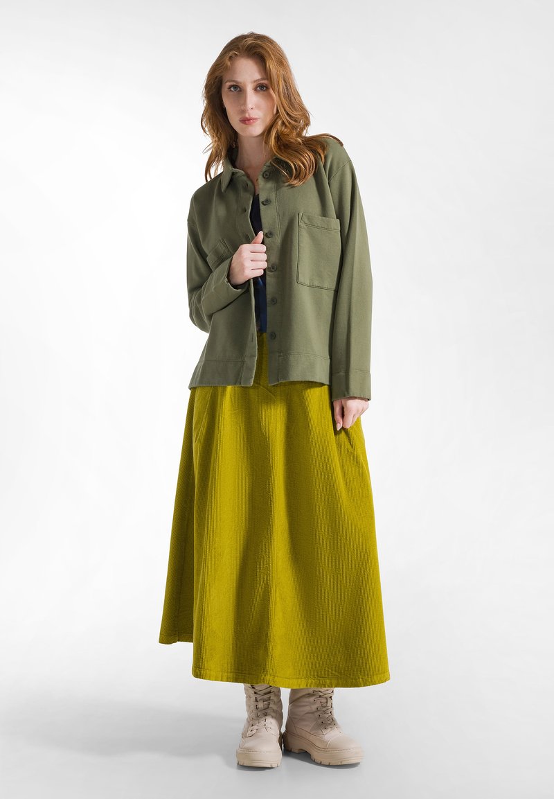 Groene katoenen shirtjasje met een borstzak, gecombineerd met een getexturede gele maxirok en beige veterschoenen.