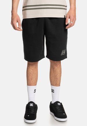 Zwarte shorts van een katoenmix, met een rechte snit. Er staat een wit logo op het linkerbeen. Gecombineerd met zwarte sneakers en witte sokken.