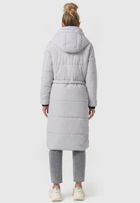 Ljust grå puffjacka med huva, lång längd, bälte i midjan, kviltad design och svarta Manschetter. Bärs över grå byxor och vita sneakers.