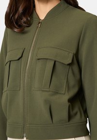 Olivgrüne Bomberjacke aus einem strukturierten Stoff. Verfügt über einen Reißverschluss, gerippte Bündchen und zwei aufgesetzte Fronttaschen.