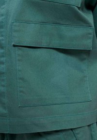 Türkiser Blazer mit zwei großen aufgesetzten Taschen, hergestellt aus einem strukturierten Stoff mit sichtbaren Nähten, die die Taschen und Nähte umreißen.