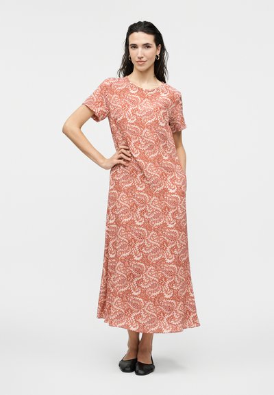 Γυναίκα με σκούρα μαλλιά, φορώντας κόκκινο φόρεμα με κοντά μανίκια και μήκος μέχρι τους αστραγάλους, με λευκά σχέδια παϊέτες, και μαύρες πλατφόρμες, στέκεται με το χέρι στη μέση.