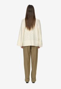 Kvinna som står med ryggen mot kameran, iförd en vit stickad tröja och lösa bruna byxor med svarta skor mot en vit bakgrund.