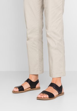Fødder iført sorte sandaler med åbne tæer og ankelstropper, kombineret med lyse beige, korte bukser på en lys grå gulvbaggrund.
