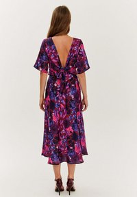 Kwiatowa sukienka z głębokim dekoltem na plecach, falbaniastymi rękawami i paskiem do związania. Tkanina ma purpurowy i różowy wzór kwiatowy na ciemnym tle.