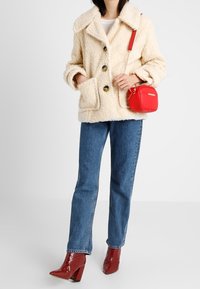 Beżowa kurtka z futra syntetycznego z kołnierzem, przednimi kieszeniami i dużymi guzikami. Połączona z niebieskimi dżinsami i czerwonymi, lakierowanymi botkami za kostkę. Czerwona torba na ramię.