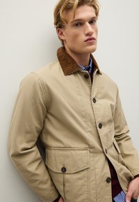 Giacca in tela beige con colletto in velluto a coste marrone, bottoni frontali e due tasche laterali. Strato interno visibile con una camicia a righe blu.