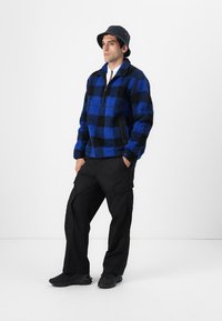 Blå og sort ternet fleecejakke med lynlås, båret med sorte cargo-bukser og en sort bucket hat. Struktureret, afslappet pasform.