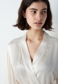 Robe in raso color crema con scollatura a forma di V e maniche lunghe, caratterizzata da una texture liscia e una leggera lucentezza. Senza motivi o hardware.