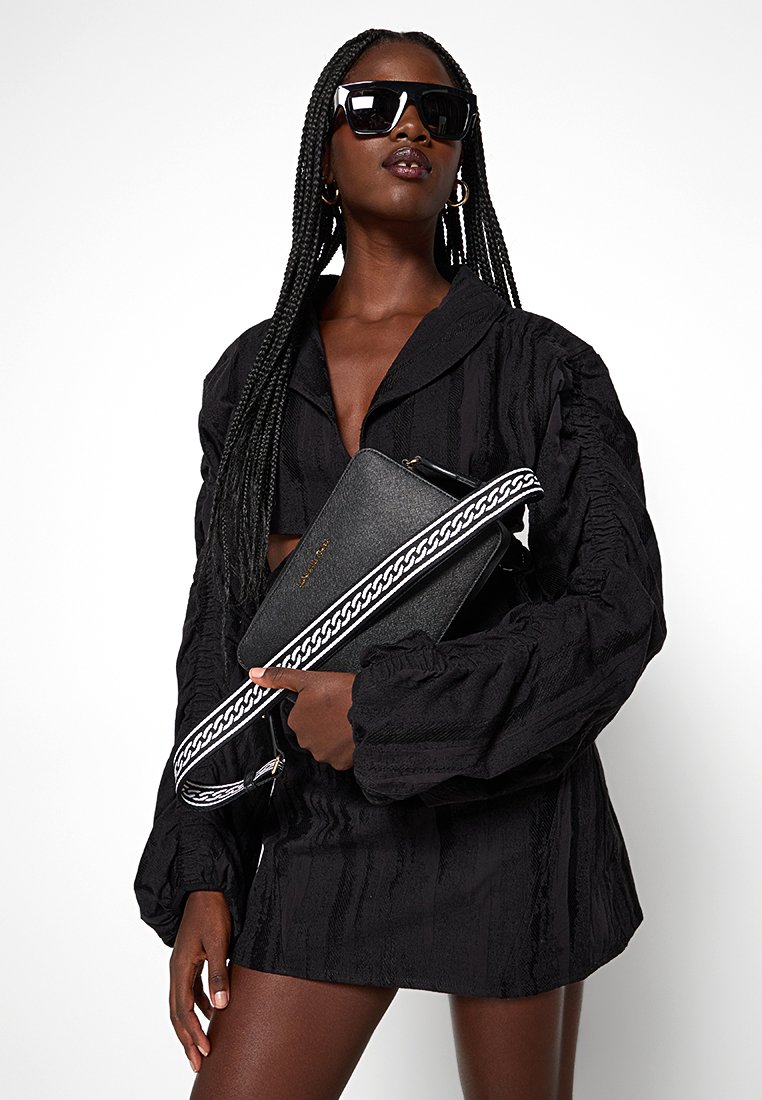 Czarna torebka na ramię o fakturowanym wykończeniu, z długim paskiem w biały wzór łańcucha. Modelka ma na sobie czarną oversize'ową koszulę i okulary przeciwsłoneczne.