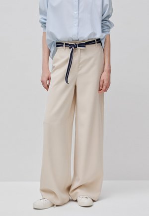 Donna che indossa pantaloni beige a gamba larga con cintura annodata a righe blu navy e bianco, camicia azzurra abbottonata e scarpe da ginnastica bianche.