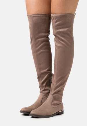 Paire de bottes taupe en daim au-dessus du genou avec fermetures éclair latérales et talons bas portées sur des jambes nues sur fond blanc.