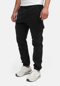 Black cargo pants with elastic waistband, drawstring, side pockets, and tapered fit. Worn with white and gray sneakers for contrast.