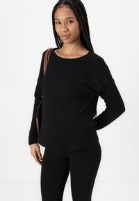 Svart langermet topp med en teksturert diamantmønster, rund hals og avslappet passform. Kombinert med matchende svarte leggings og en brun skulderveske.