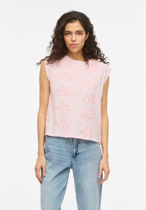 Femeie cu păr creț purtând un top fără mâneci, roz deschis, brodat cu flori, și blugi albaștri deschis, stând într-un cadru alb.