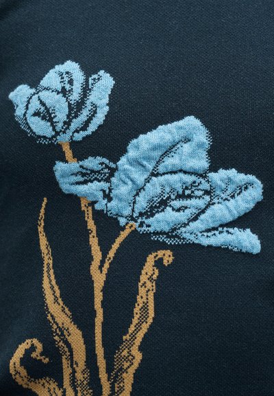 Μπλε και μπεζ κεντήματα με δύο λουλούδια και φύλλα σε σκουρόχρωμο ύφασμα, που δείχνει λεπτομερή υφή πλεξίματος.