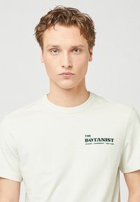 Lichtgekleurde t-shirt met korte mouwen, gemaakt van katoen. Functies "THE BOTANIST" logo afgedrukt in groen aan de bovenkant links.
