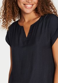 Blusa blu navy con scollatura a V e maniche corte, caratterizzata da un delicato motivo a pois e un tessuto leggero, che offre una superficie liscia.