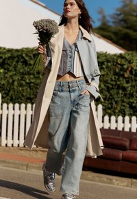 Trench coat beige ligero sobre un top corto de mezclilla azul y jeans de pierna ancha azul claro. Zapatillas plateadas y un ramo de lavanda.