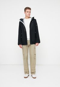 Noor mees seisab beežides cargo pükstes, valges särgis, mustas kapuutsiga jakis ja valgetes tossudes, lihtsa valge tausta ees.