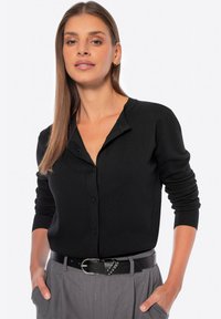 Cardigan nero a maglia con chiusura a bottoni frontale, polsini a coste e colletto poco profondo. Indossato sopra pantaloni grigi, abbinato a una cintura nera con borchie.