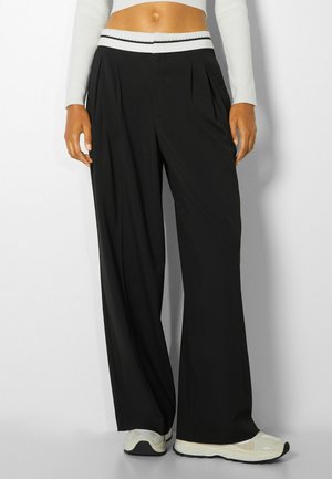 Kobieta w czarnych, szerokich spodniach z białym pasem, białym, krótkim topie z długim rękawem i białych trampkach stoi na białym tle.