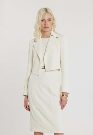 Donna con capelli biondi che indossa un vestito bianco aderente con un blazer corto abbinato e accessori dorati su uno sfondo semplice.