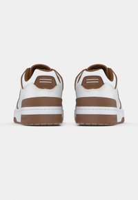 Ζευγάρι λευκά και καφέ αθλητικά παπούτσια Lacoste, εμφανιζόμενα από πίσω σε απλό λευκό φόντο.