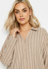 Camicia button-up beige con sottili righe verticali nere, colletto e maniche lunghe. Il materiale appare tessurizzato e leggermente stropicciato.