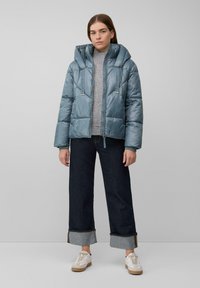 Veste matelassée bleu clair avec capuche, fermeture éclair à l'avant et détails à cordon, portée sur un pull gris, associée à un jean foncé et des baskets blanches.