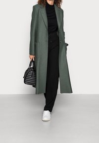 Abrigo largo verde hecho de tela texturizada, con un diseño de doble botonadura, grandes bolsillos, y combinado con pantalones negros, zapatillas blancas y un bolso negro.