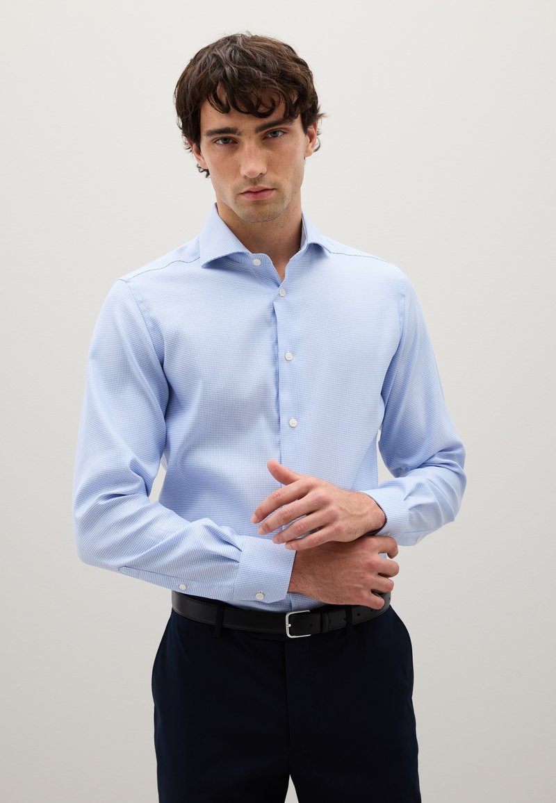 Giovane uomo che sistema il polsino di una camicia da uomo azzurro chiaro, indossando una cintura nera e pantaloni scuri, in piedi davanti a uno sfondo chiaro uniforme.