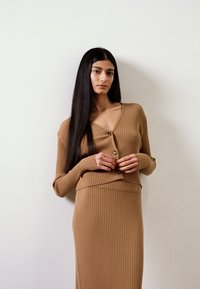 Conjunto de malha canelada em tom bege, composto por saia ajustada e um cardigan de decote em V com botões. Textura suave e design justo.