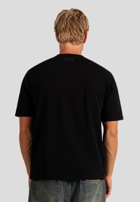Černé bavlněné tričko s krátkými rukávy a kulatým výstřihem. Má čtvercovou nášivku na zadní straně výstřihu. Jednoduchý, čistý design.