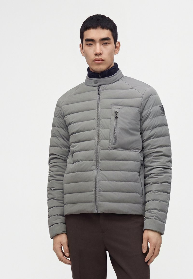 Jeune homme portant une veste matelassée gris clair avec des poches zippées et un pantalon foncé, debout devant un fond uni.