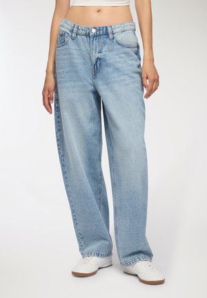 Jeans in denim azzurro chiaro, oversize, con vita alta, cinque tasche e orli risvoltati, abbinati a sneaker bianche su uno sfondo neutro.