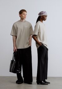 Două manechine stau una lângă alta. Prima poartă un tricou supradimensionat bej și pantaloni largi negri, ținând o geantă tote neagră. A doua poartă un tricou similar și un batic cu model.