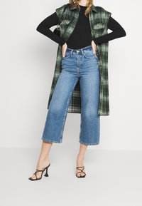 Överdimensionerad grönrutig skjortjacka över en svart ribbad långärmad topp, kombinerad med vida blå denimjeans och svarta remmar med hög klack.